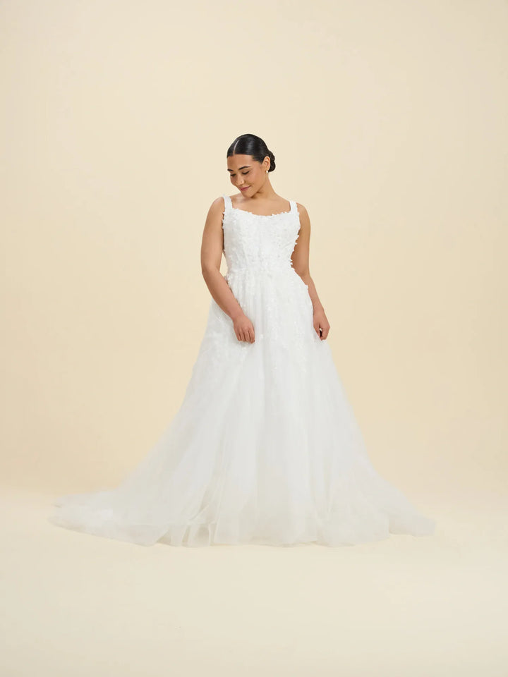 A woman modeling a white sleeveless wedding gown with a fitted bodice, square neckline, and a full layered tulle skirt against a plain warm beige studio background.