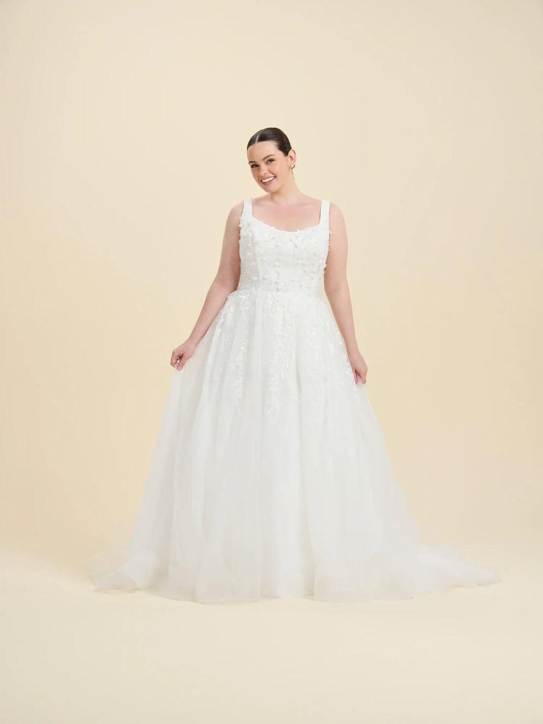 A woman modeling a white sleeveless wedding gown with a fitted lace bodice and a full tulle skirt, standing against a plain warm beige studio background.