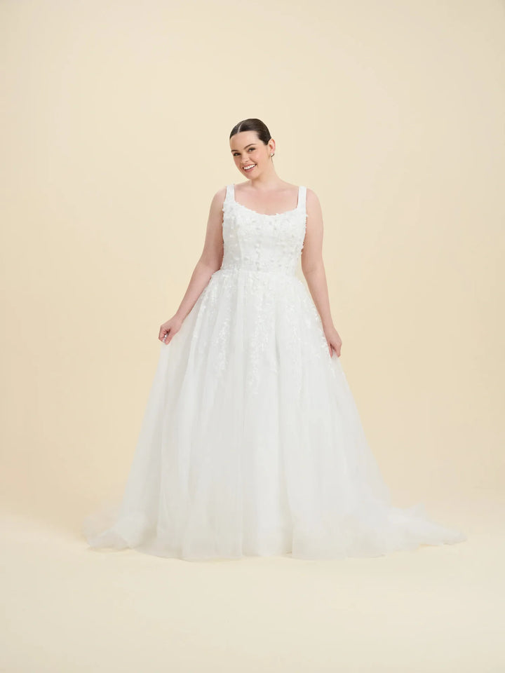 A woman modeling a white sleeveless wedding gown with a fitted lace bodice and a full tulle skirt, standing against a plain warm beige studio background.
