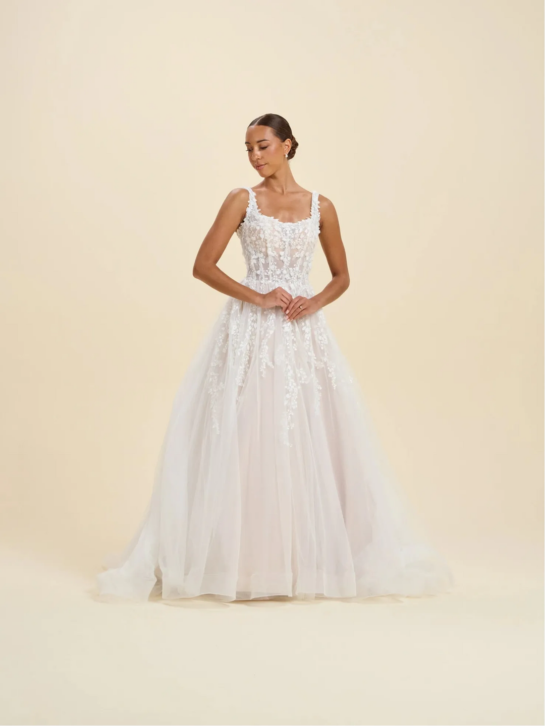 A woman modeling a white sleeveless wedding gown with a fitted lace bodice and a full tulle skirt, standing against a plain warm beige studio background.