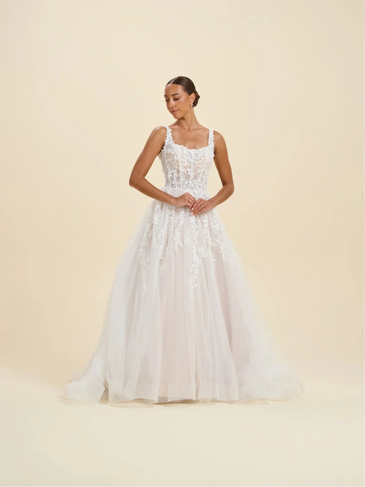 A woman modeling a white sleeveless wedding gown with a fitted lace bodice and a full tulle skirt, standing against a plain warm beige studio background.