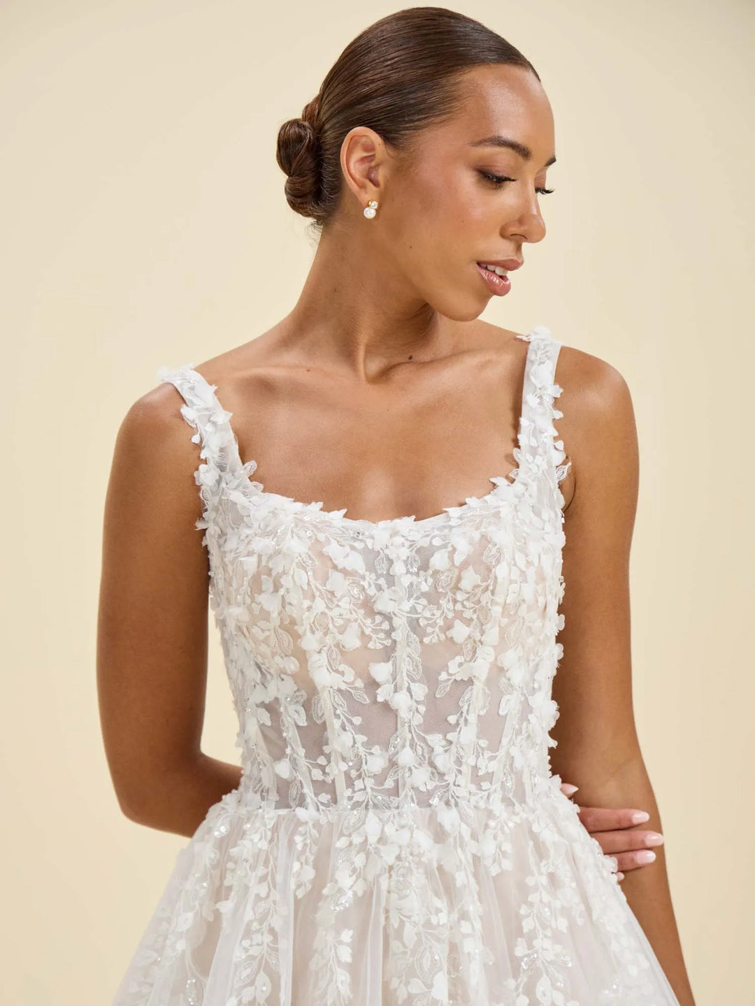 A woman wearing a white sleeveless lace dress with a fitted bodice and flared tulle skirt, photographed against a plain warm beige studio background.