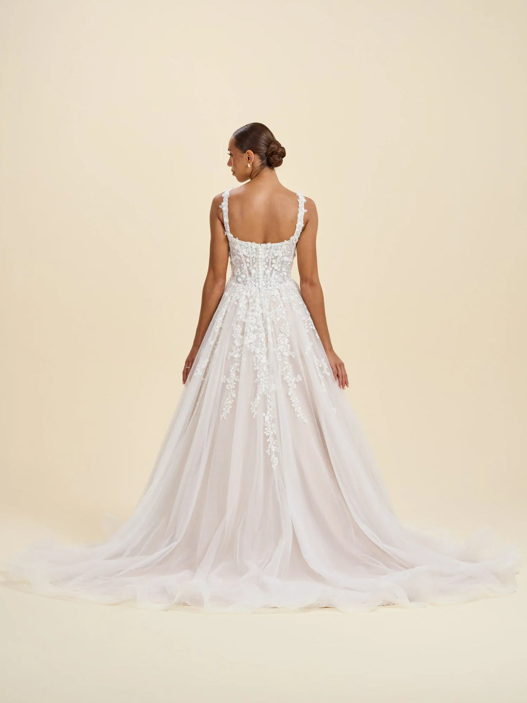 A woman modeling a white bridal gown from the back, showing a fitted lace bodice, thin straps, and a full tulle skirt with a long train against a warm cream studio background.