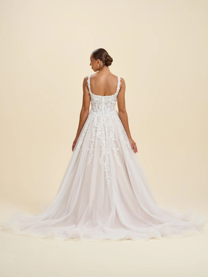 A woman modeling a white bridal gown from the back, showing a fitted lace bodice, thin straps, and a full tulle skirt with a long train against a warm cream studio background.