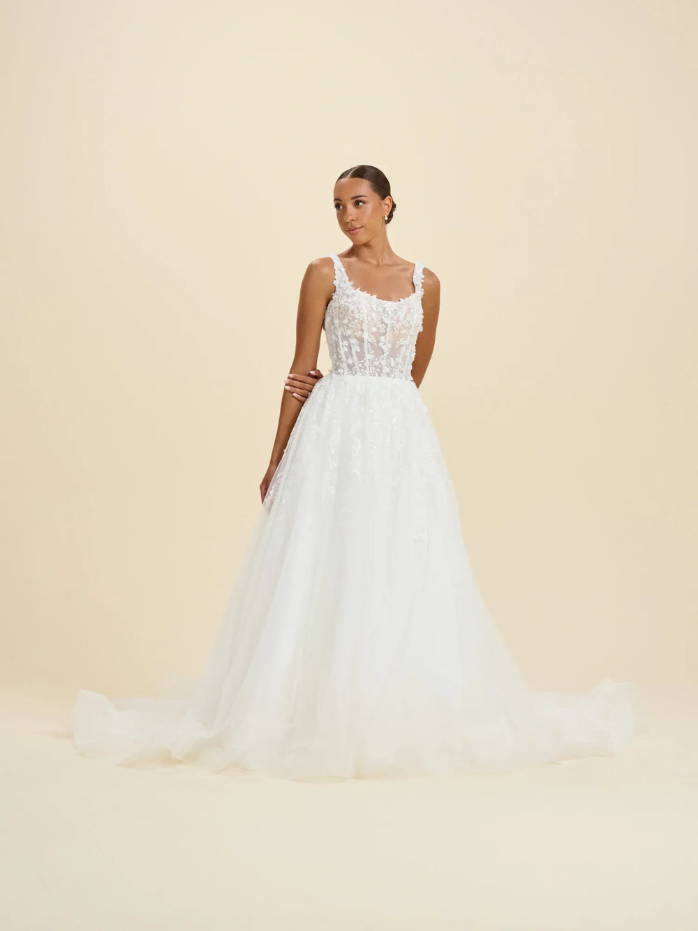 A woman modeling a white sleeveless wedding gown with a lace bodice, thin straps, and a full tulle skirt against a plain beige studio background.