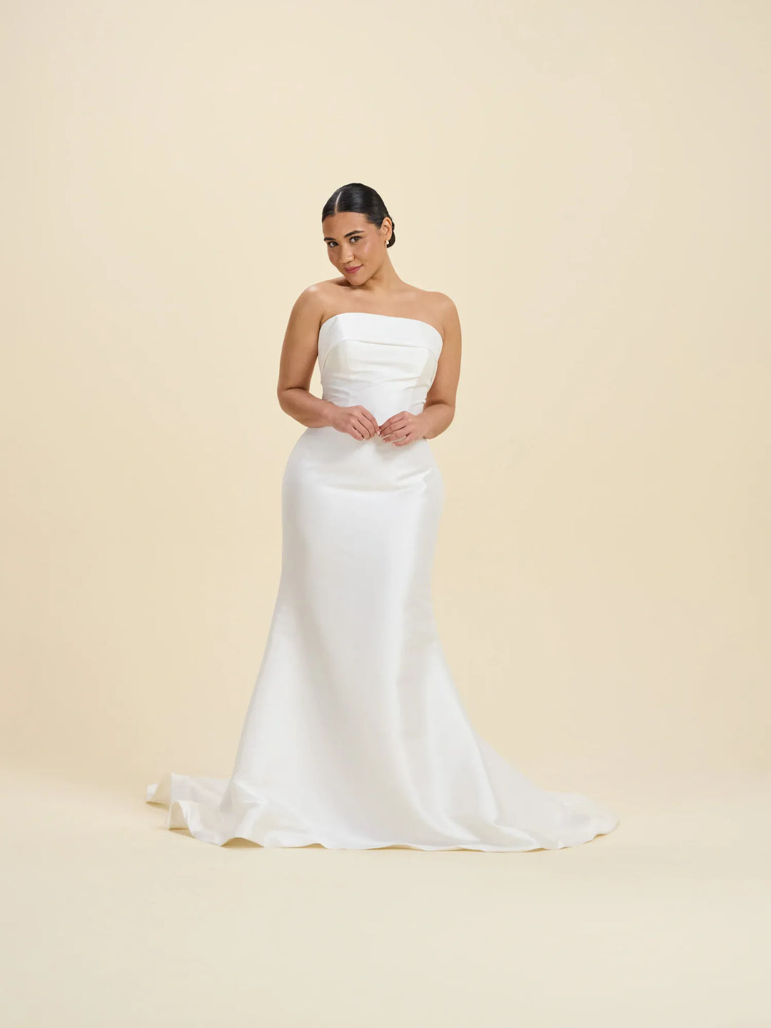 A woman modeling a strapless white satin wedding gown with a fitted bodice and a slight train, standing against a plain warm beige studio background.