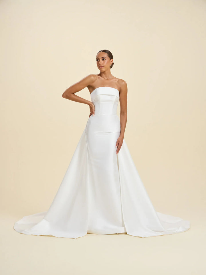 A woman modeling a strapless white wedding gown with a fitted bodice and a full satin train, posed against a plain warm beige studio background.