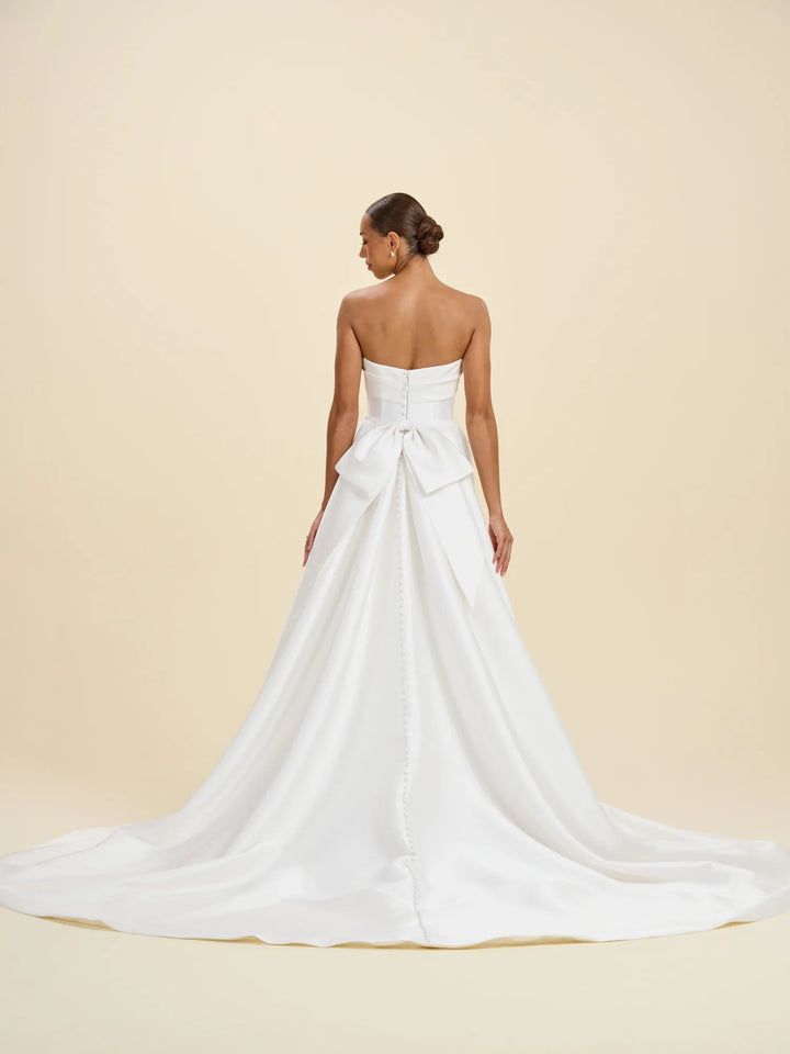 A woman modeling a strapless white wedding gown from the back, showing a structured bodice, large bow detail at the waist, and a full satin train against a warm beige studio background.