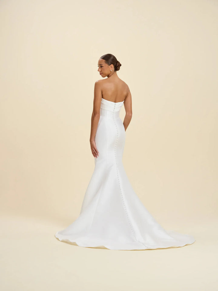 A woman modeling a strapless white mermaid-style wedding gown from the back, standing against a plain warm beige studio background.