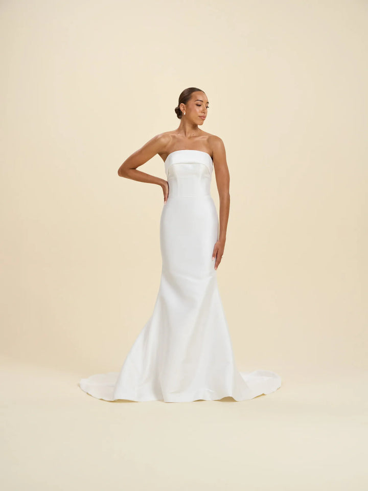 A woman modeling a strapless white mermaid-style wedding gown with a fitted bodice and flared skirt, posed against a plain warm beige studio background.