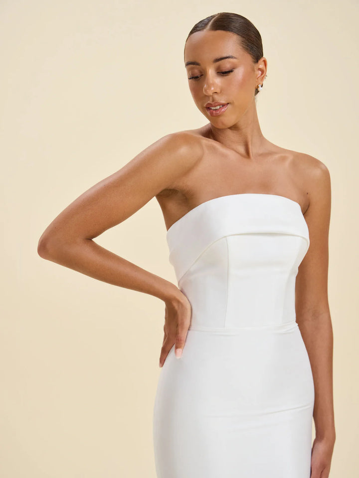 A woman modeling a strapless white fitted dress against a warm beige studio background.