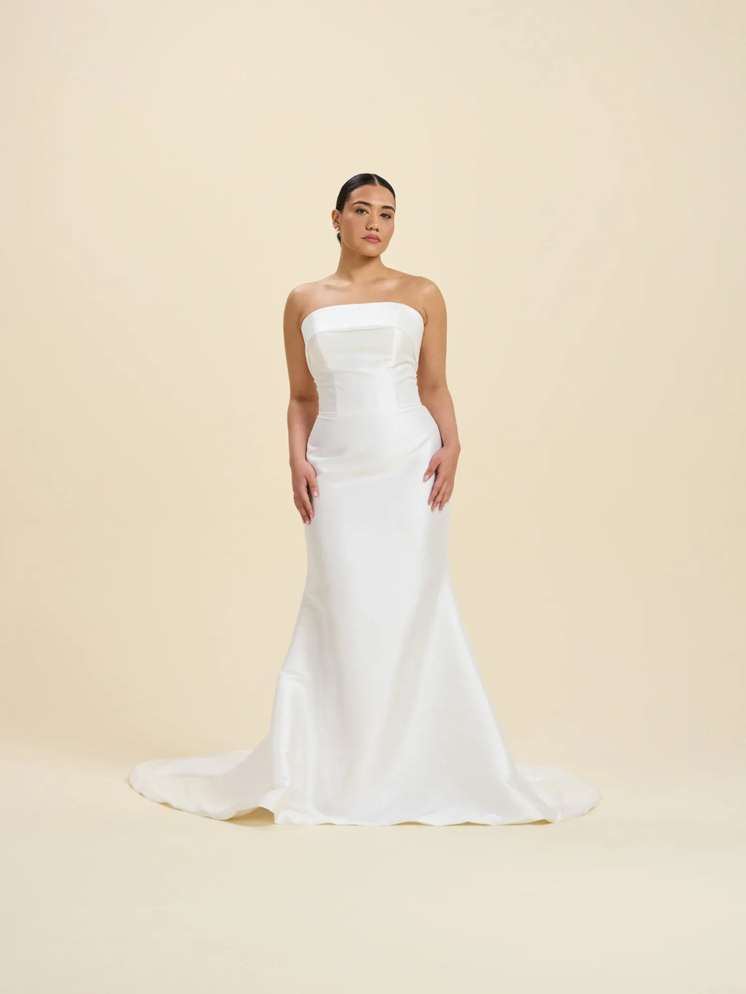 A woman modeling a strapless white mermaid-style wedding gown with a fitted bodice and slight train, standing against a plain warm beige studio background.