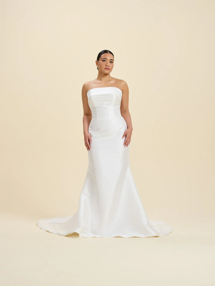 A woman modeling a strapless white mermaid-style wedding gown with a fitted bodice and slight train, standing against a plain warm beige studio background.