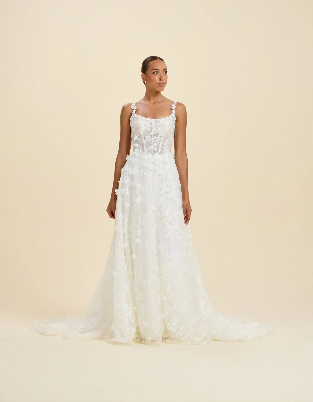 A woman modeling a sleeveless white wedding gown with a fitted lace bodice and a full, textured skirt with a slight train, photographed against a plain warm beige studio background.