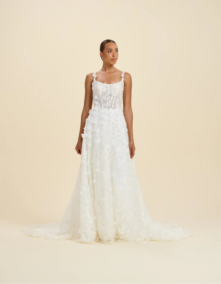 A woman modeling a sleeveless white wedding gown with a fitted lace bodice and a full, textured skirt with a slight train, photographed against a plain warm beige studio background.