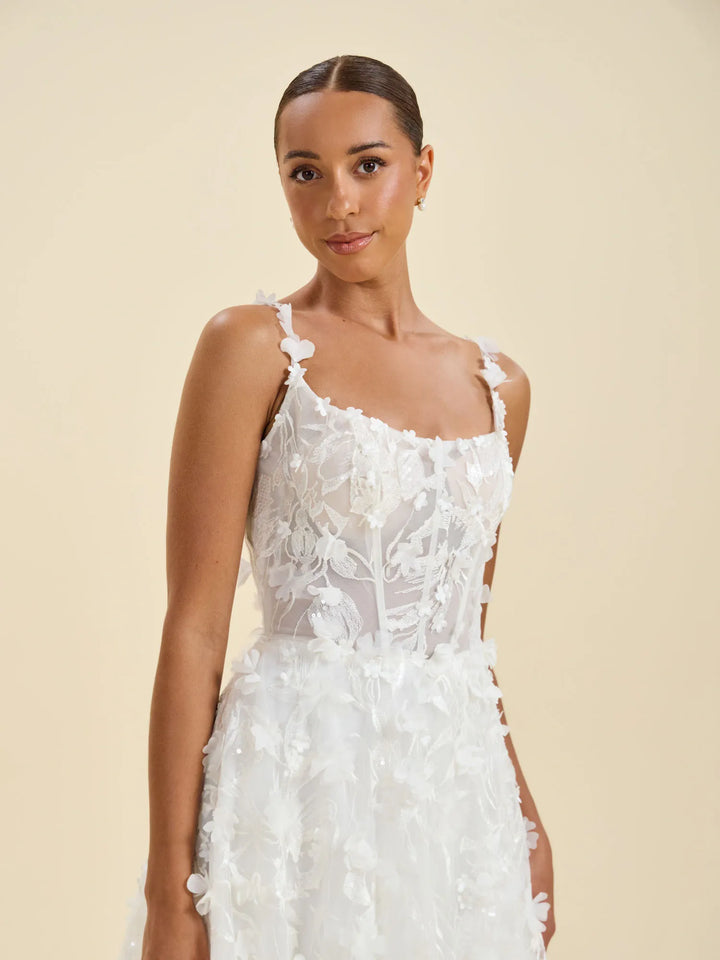 A woman modeling a white sleeveless wedding dress with floral lace applique and 3D flower details against a solid beige background.
