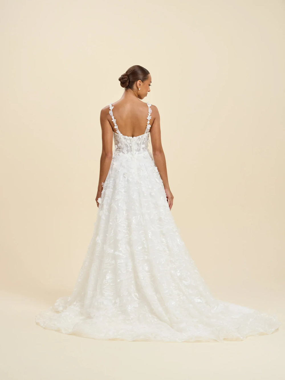 A woman modeling a white lace wedding gown from the back against a warm beige studio background.