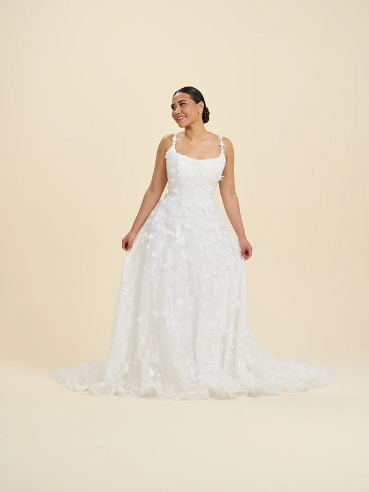 A woman modeling a white sleeveless wedding gown with thin straps, a fitted bodice, and a full lace-embroidered skirt with a slight train, photographed against a plain warm cream studio background.