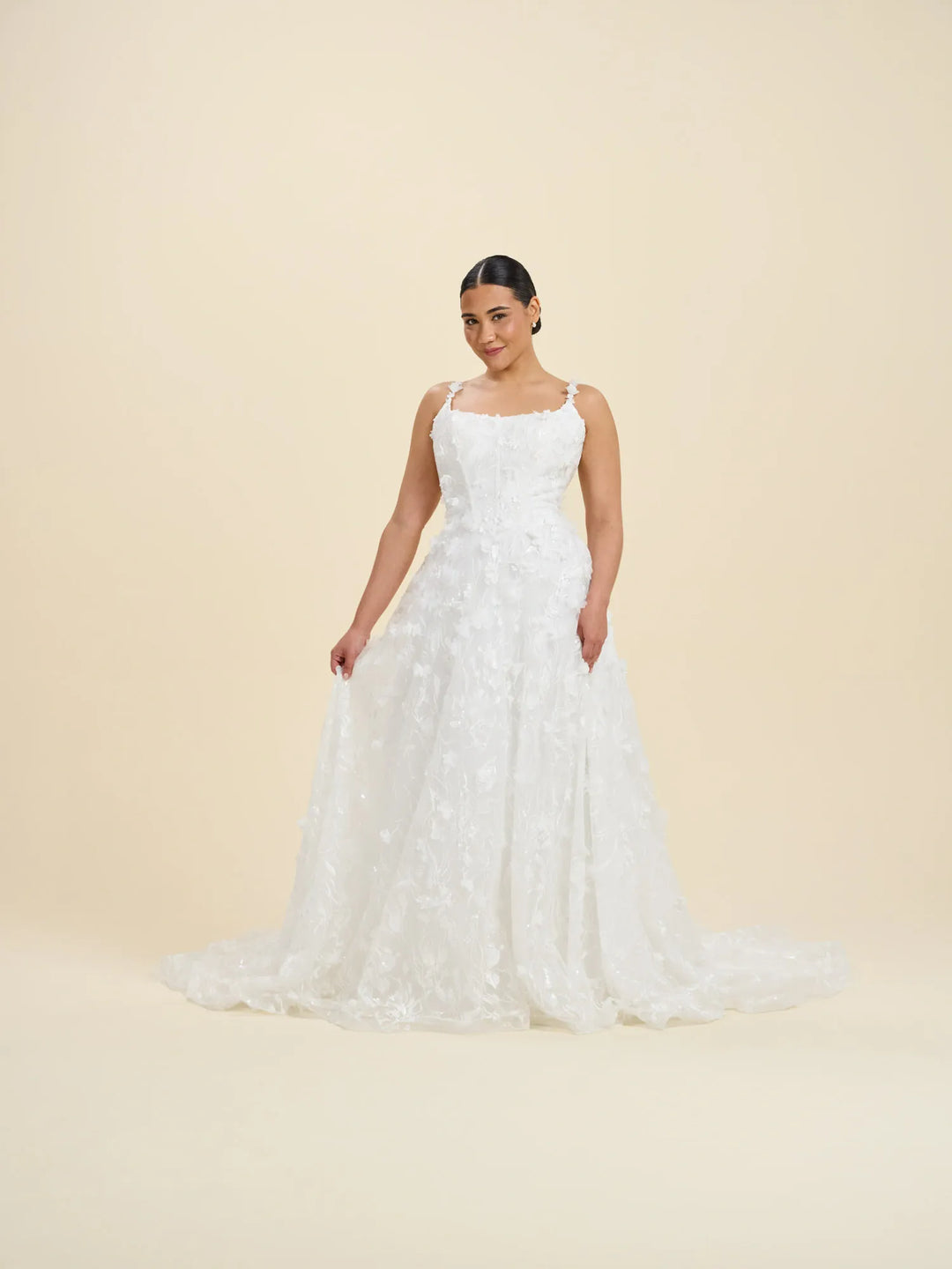 A woman modeling a white sleeveless wedding gown with a fitted bodice, thin straps, and a full lace-embroidered A-line skirt against a plain beige studio background.