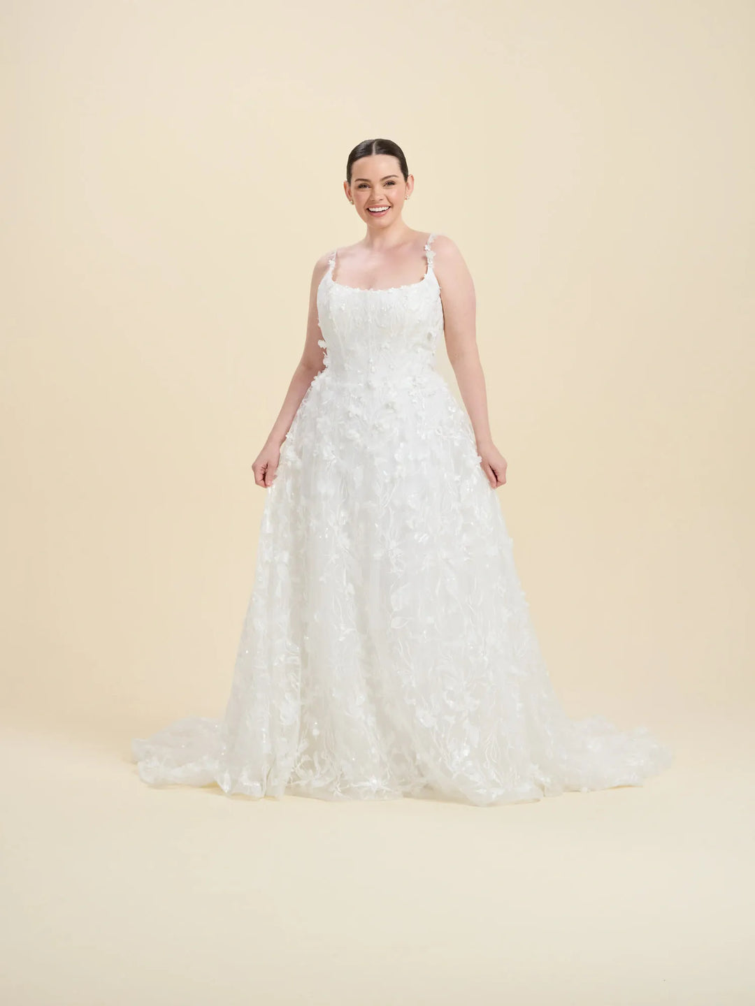 A woman modeling a white sleeveless A-line wedding gown with a fitted bodice and full lace-trimmed skirt, standing against a plain warm beige studio background.