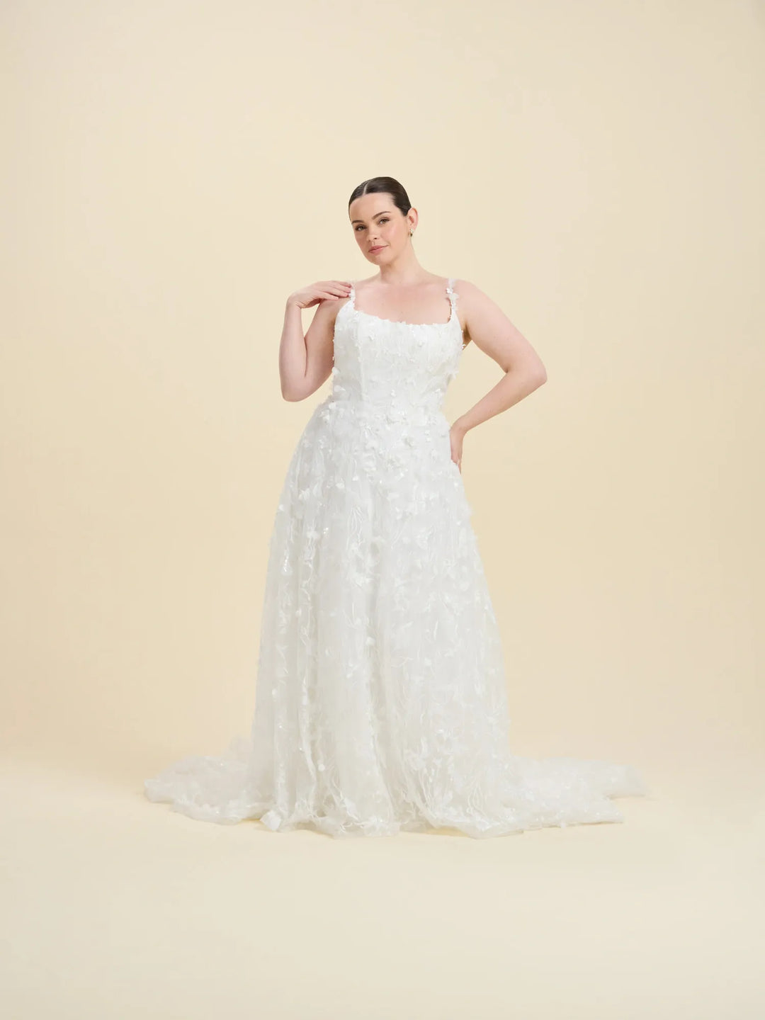 A woman modeling a sleeveless white wedding gown with a fitted bodice, thin straps, and a full lace-embroidered skirt with a slight train, posed against a solid light beige studio backdrop.