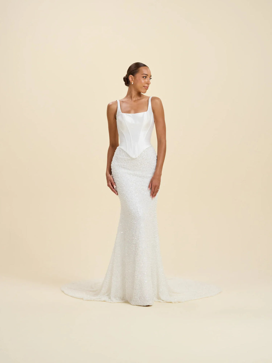 A woman modeling a sleeveless white wedding gown with a structured bodice, fitted sequined skirt, and a short train against a plain warm beige studio background.