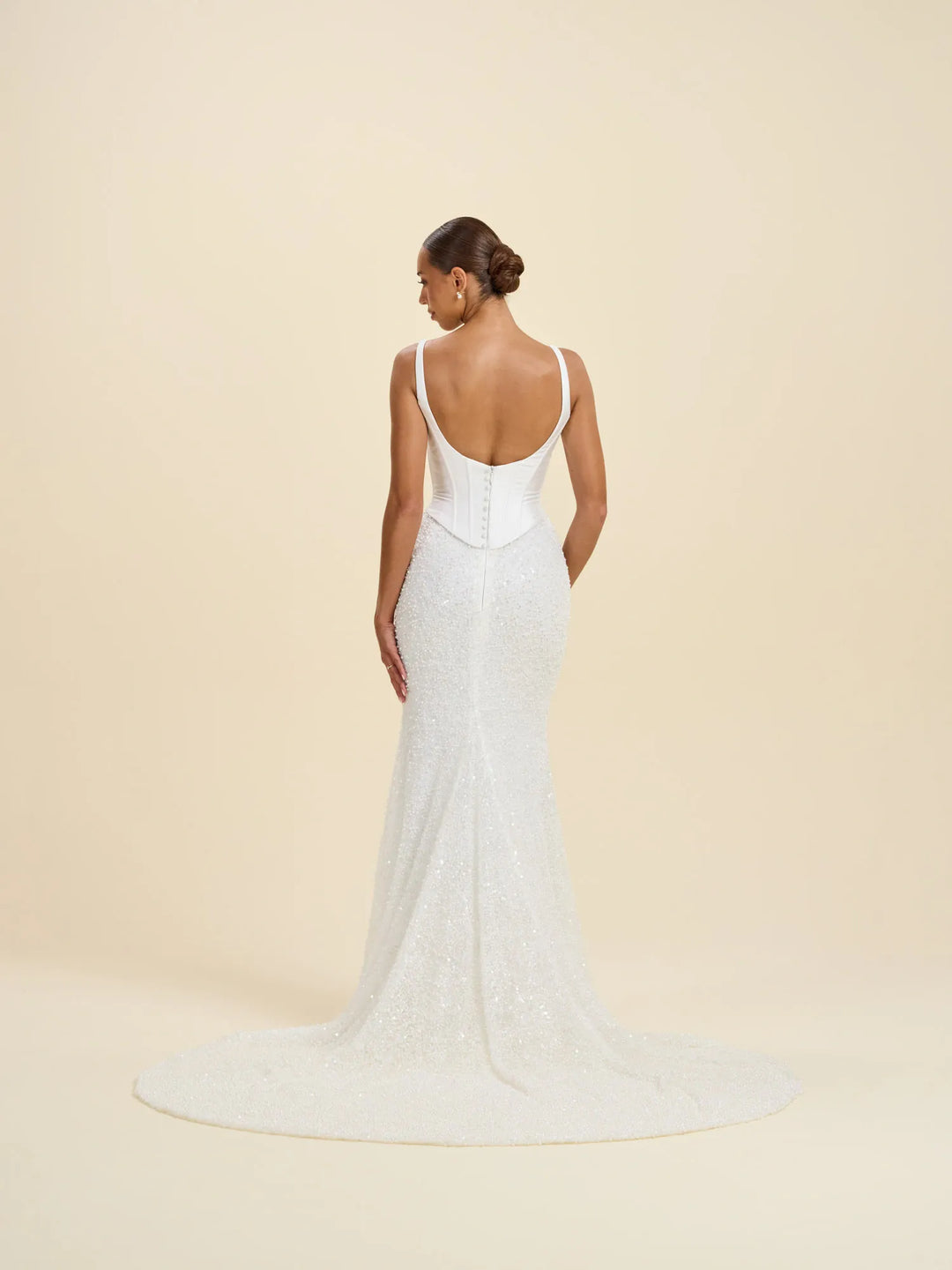 A woman modeling a white sleeveless wedding gown from the back, showing a deep open back, fitted silhouette, and a short train, against a plain warm beige studio background.