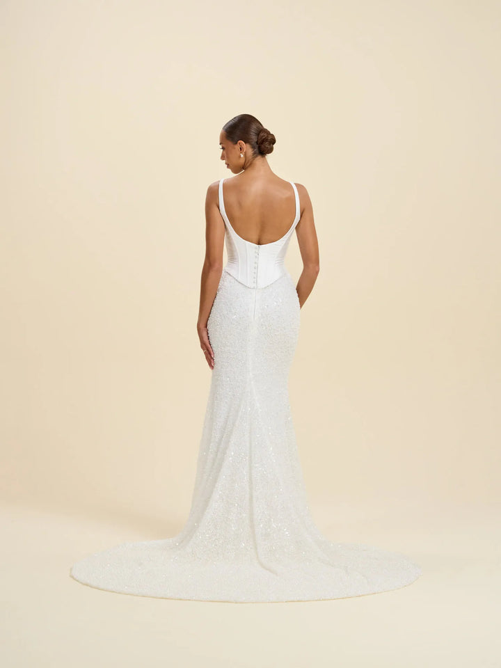A woman modeling a white sleeveless wedding gown from the back, showing a deep open back, fitted silhouette, and a short train, against a plain warm beige studio background.