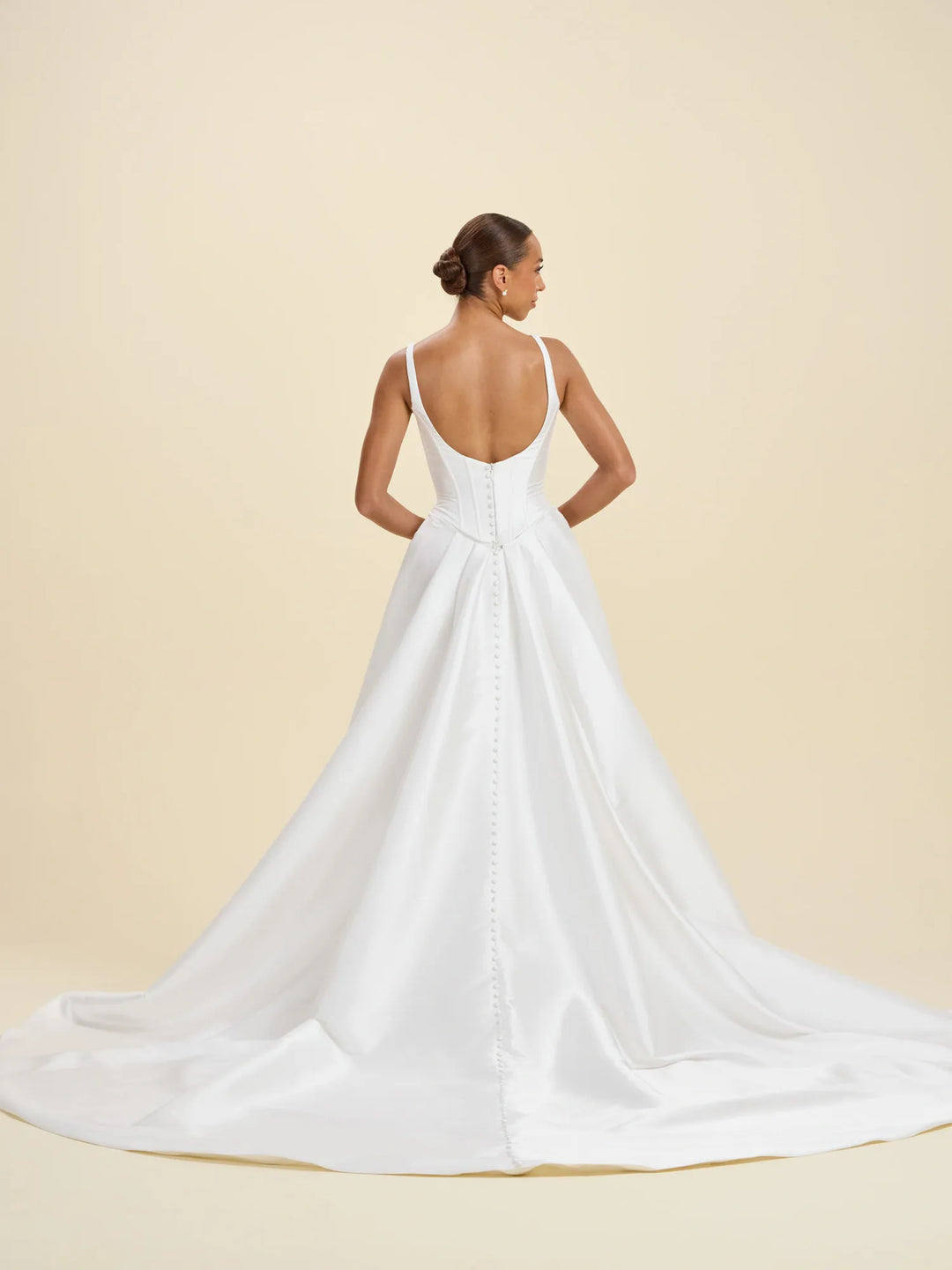 A woman modeling a white bridal gown from the back, showing a low open back, thin straps, and a full satin train against a warm beige studio background.