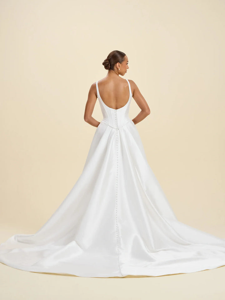 A woman modeling a white bridal gown from the back, showing a low open back, thin straps, and a full satin train against a warm beige studio background.