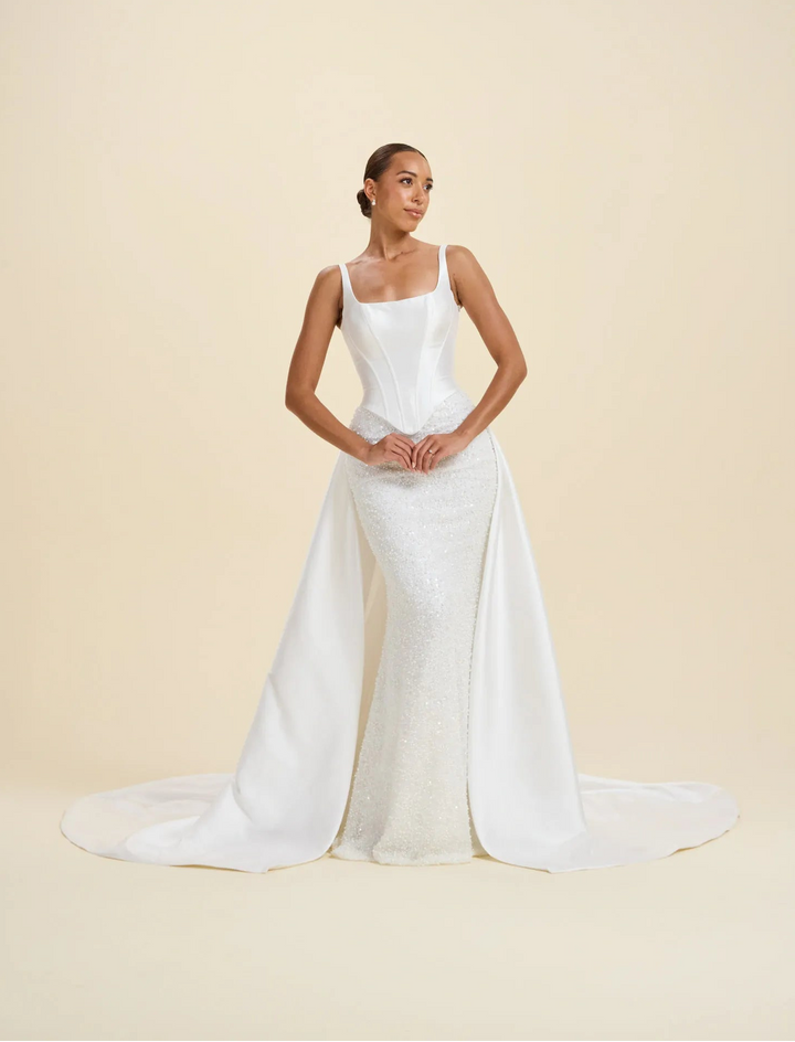 A woman modeling a white sleeveless wedding gown with a fitted bodice, textured skirt, and long train against a plain beige studio background.