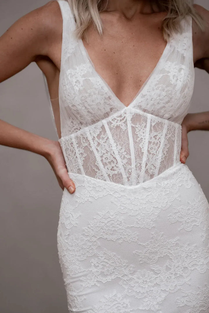 White lace wedding dress with a deep V-neckline, sheer lace corset-style waist panel, and fitted silhouette, shown on a model against a plain taupe background.