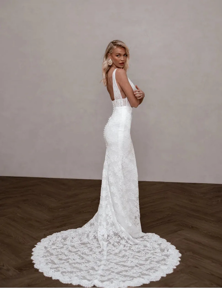 A woman modeling a white lace wedding gown from the back, standing on a dark wood floor against a textured beige wall.