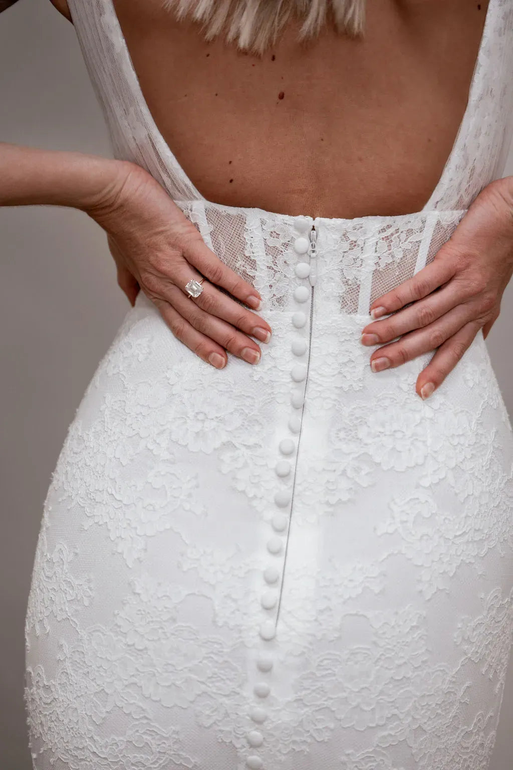 Close-up back view of a white lace bridal gown with a deep open back, decorative button detail down the center, and a model's hands resting on the hips.