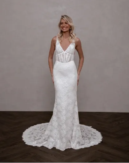 A woman modeling a white lace wedding gown with a deep V-neckline, sheer lace bodice, and a flared lace train on a dark wood floor against a textured beige wall.