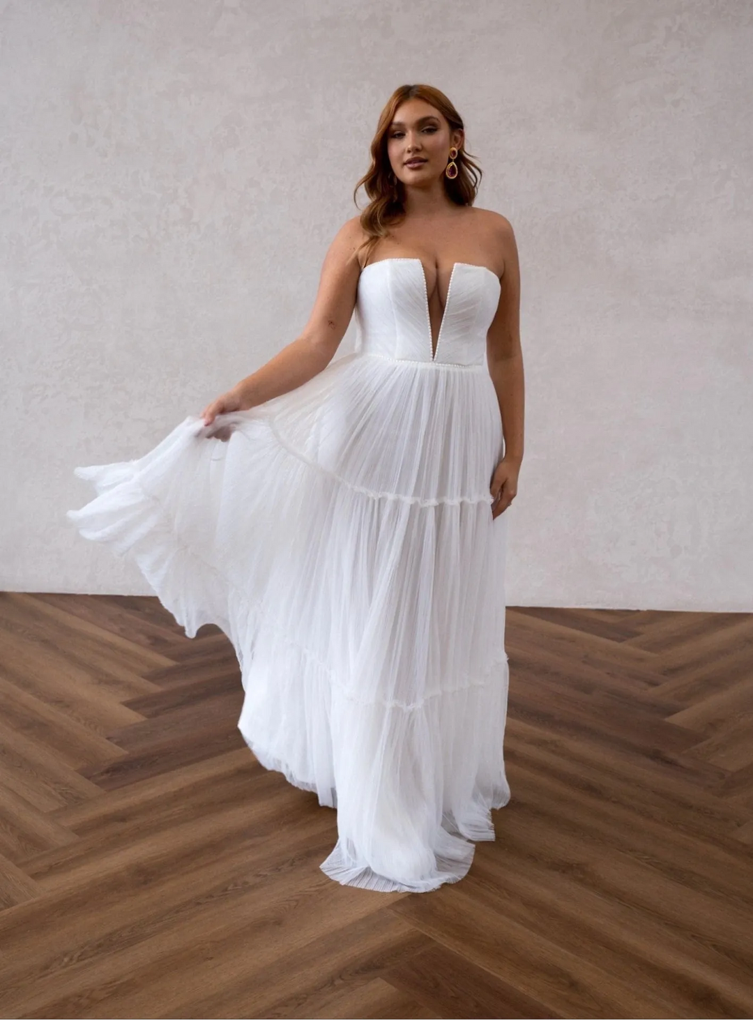 Woman in a strapless white wedding dress standing on a wooden floor with a plain wall background