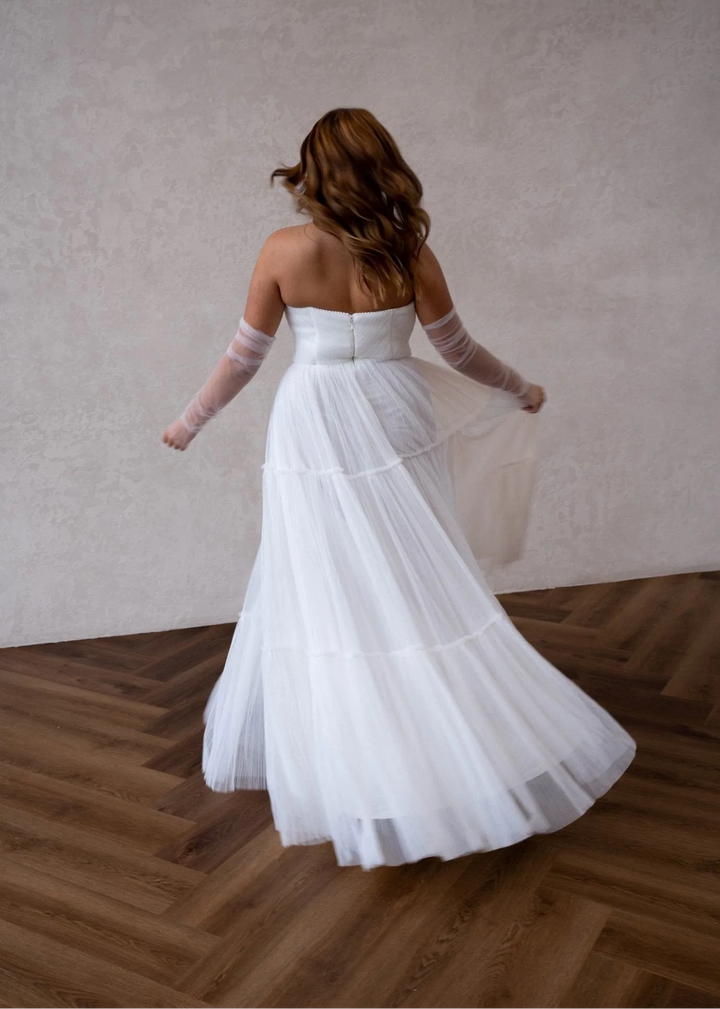 Woman in a white wedding dress with sheer sleeves standing on a wooden floor.