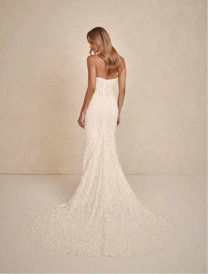 A woman wearing a strapless ivory lace wedding gown is shown from the back against a warm beige studio backdrop. The dress has a fitted silhouette with a long train.