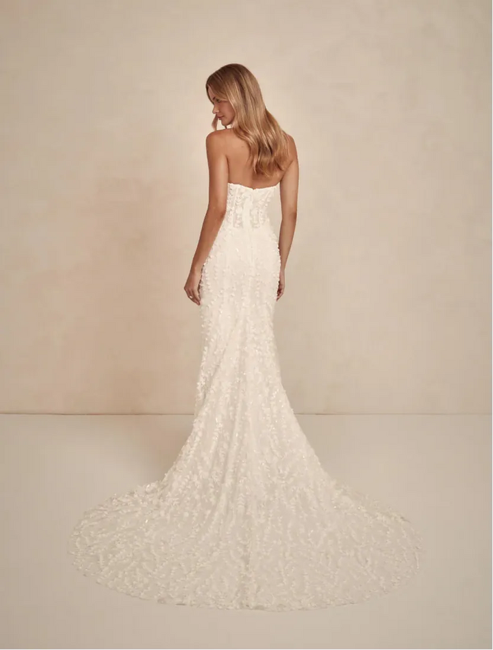 A woman wearing a strapless ivory lace wedding gown is shown from the back against a warm beige studio backdrop. The dress has a fitted silhouette with a long train.