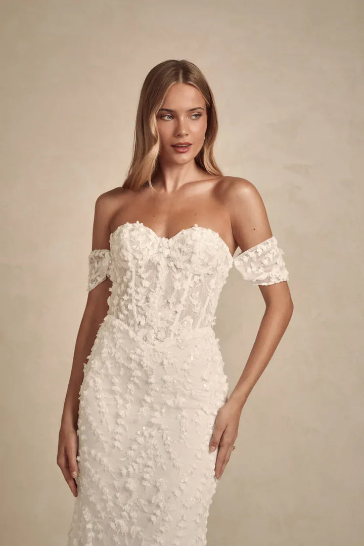 A woman modeling an off-the-shoulder white lace wedding dress with a sweetheart neckline and textured floral applique, set against a warm beige studio background.