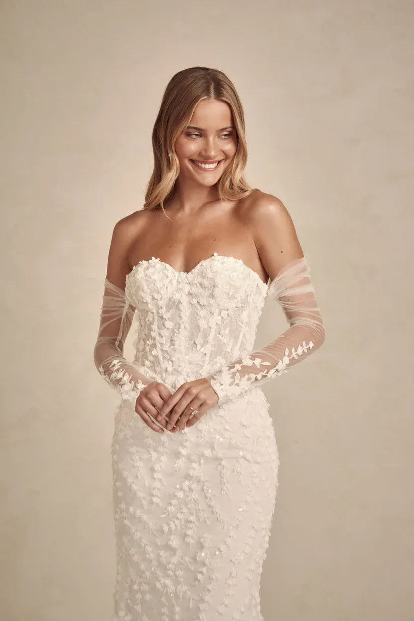A woman wearing a strapless white lace wedding gown with sheer long sleeves, standing against a warm beige studio background.