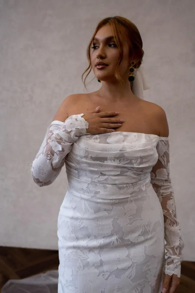 close up of made with love kennedy ivory lace sheath wedding dress with an ff the shoulder neckline and slit in the skirt
