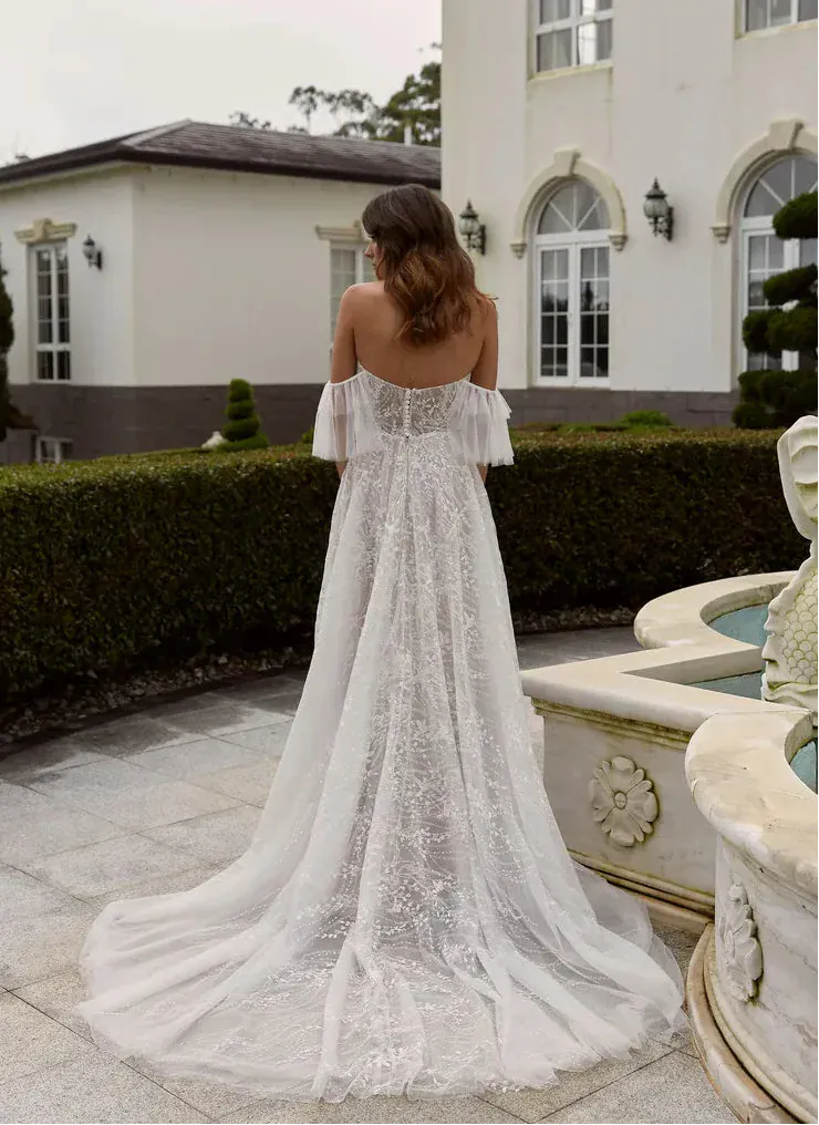 Woman in a white lace dress standing in front of a building with a fountain.