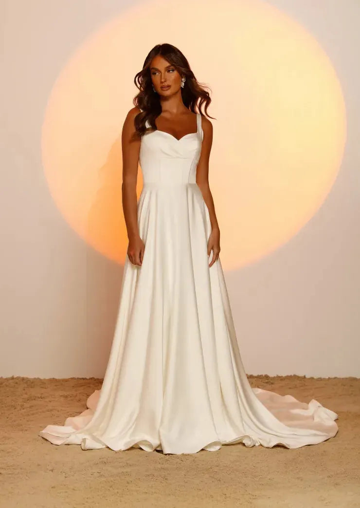 A woman modeling a sleeveless white wedding gown with a sweetheart neckline, fitted bodice, and full flowing skirt with a train, standing against a warm beige backdrop with a large soft orange spotlight behind her.