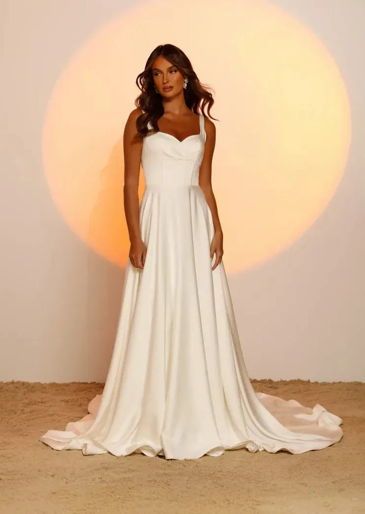 A woman modeling a sleeveless white wedding gown with a sweetheart neckline, fitted bodice, and full flowing skirt with a train, standing against a warm beige backdrop with a large soft orange spotlight behind her.