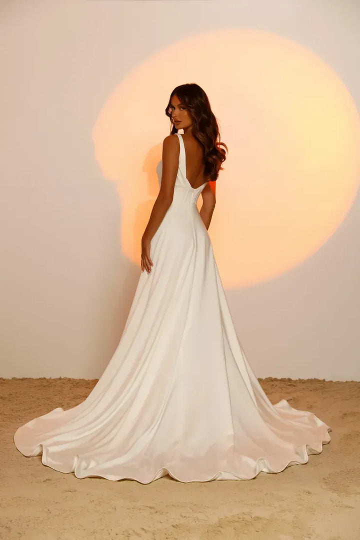A woman wearing a white sleeveless wedding gown with a deep V back and a long flowing train, standing in a studio with a warm spotlight behind her.