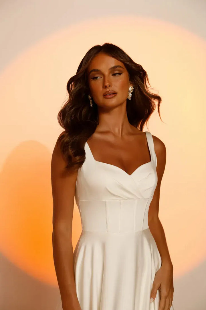 A woman modeling a white sleeveless sweetheart-neck dress with structured corset-style bodice panels and a flared skirt, posed against a warm peach and cream studio backdrop.