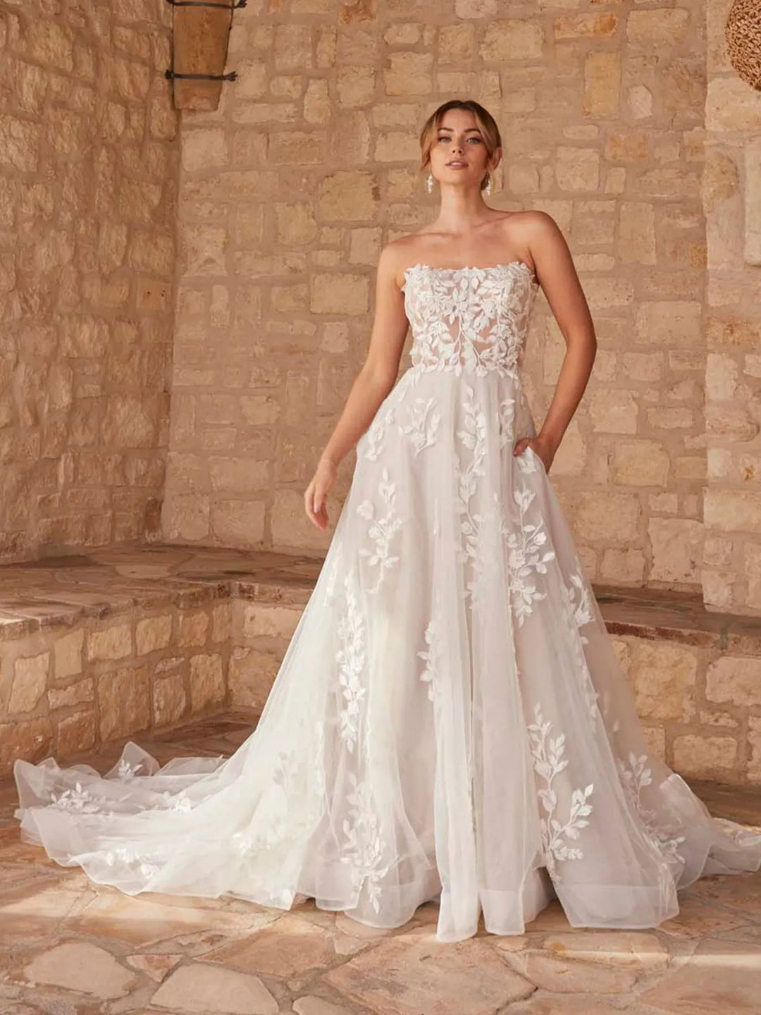 A woman wearing a strapless white wedding gown with floral lace applique and a full tulle skirt, standing in a stone-walled interior with a stone floor.