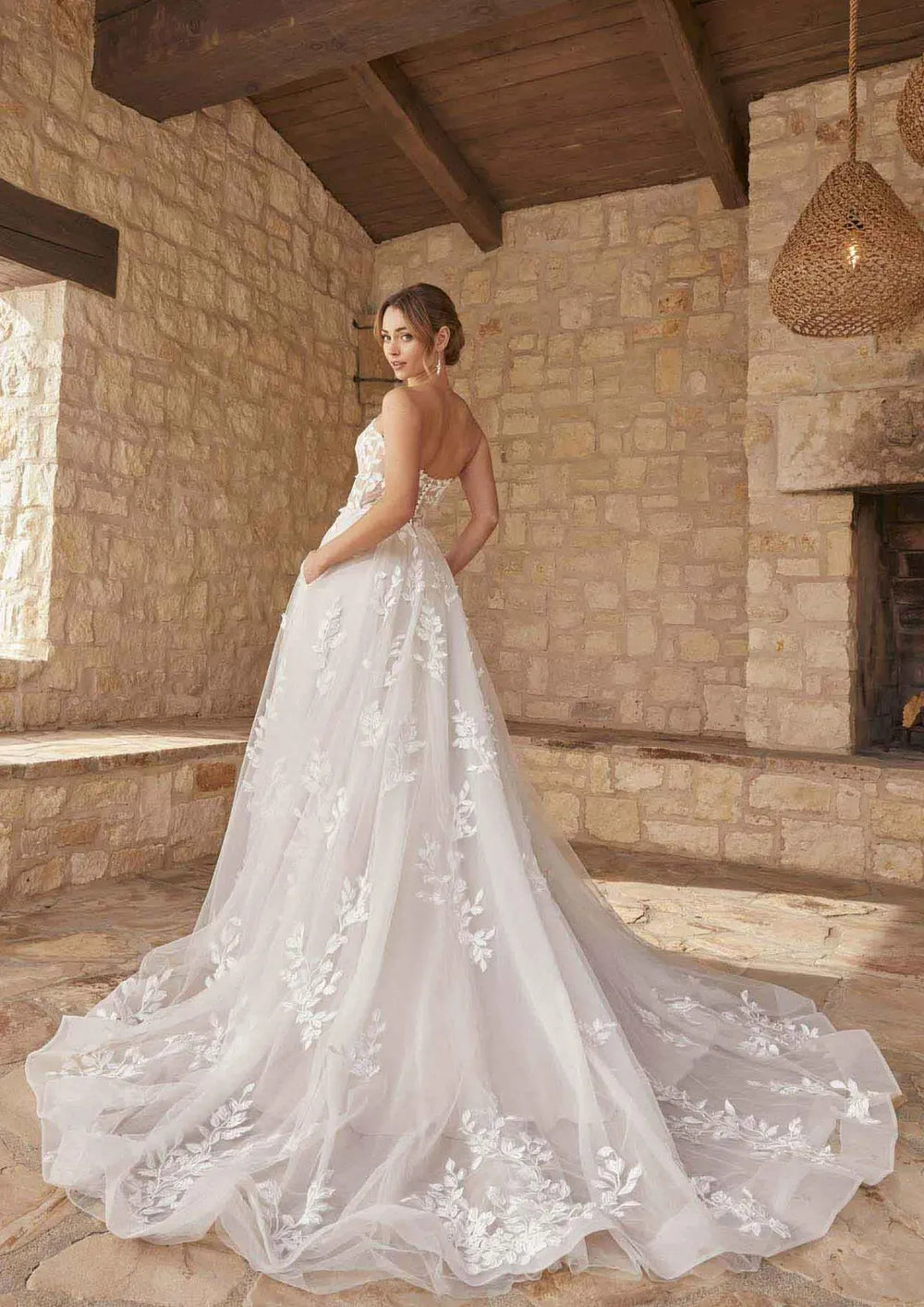 A woman wearing a white lace wedding gown stands in a rustic stone interior with wooden ceiling beams and woven pendant lights, photographed from the back to show the full train of the dress.