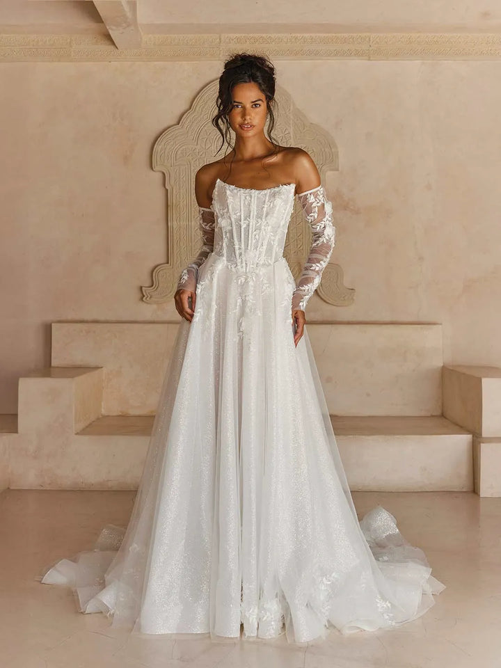 A woman modeling an off-the-shoulder white wedding gown with long sheer lace sleeves, a fitted corset-style bodice, and a full tulle skirt with a train, posed in front of a beige architectural backdrop.